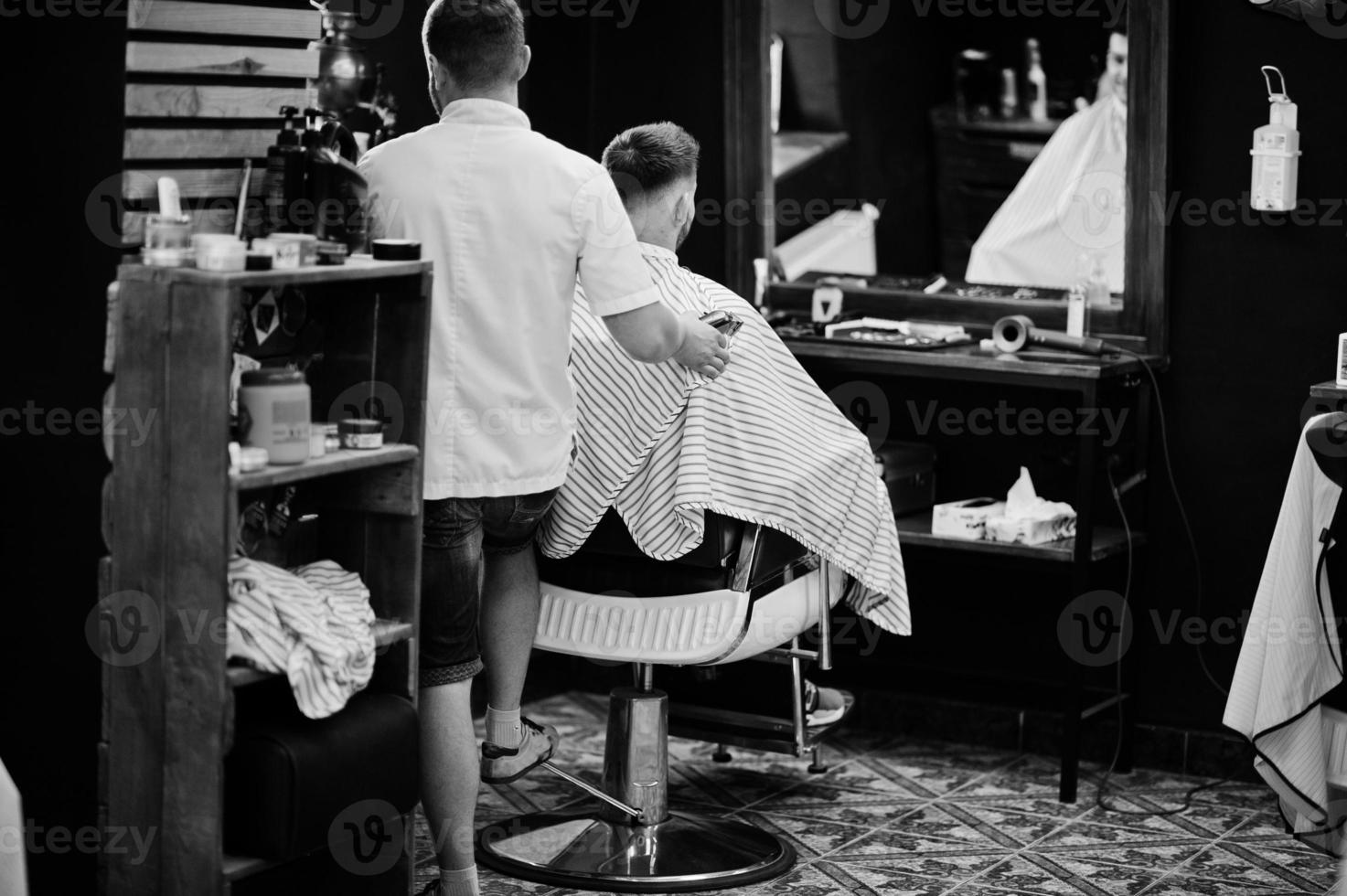 Young bearded man getting haircut by hairdresser while sitting in chair at barbershop. Barber soul. photo