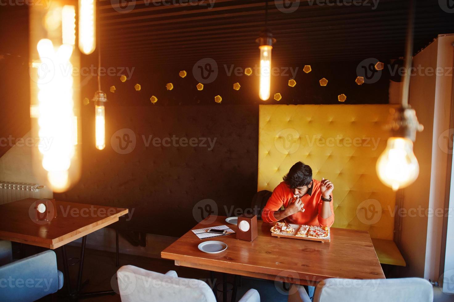 Confident young indian man in orange sweater sitting at pizzeria and eat pizza. photo