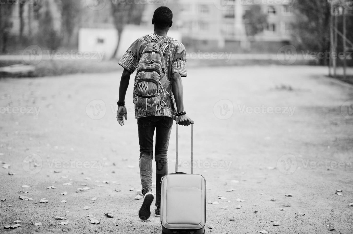 African man in africa traditional shirt on autumn park with backpack and suitcase. Emigrant traveler. photo