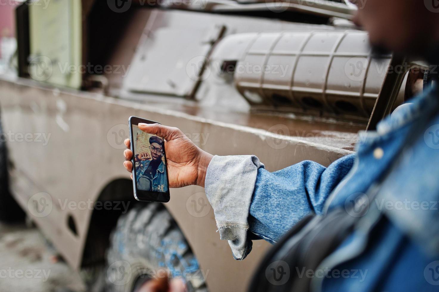 hombre afroamericano con chaqueta de jeans, boina y anteojos, fumando cigarro y posó contra un vehículo blindado militar btr, haciendo selfie por teléfono. centrarse en la pantalla del teléfono móvil. foto