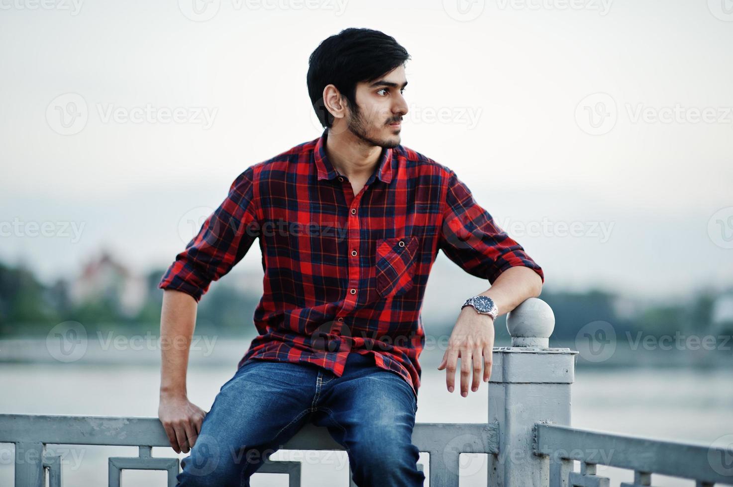 joven estudiante indio con camisa a cuadros y jeans sentado en pasamanos contra el lago. foto