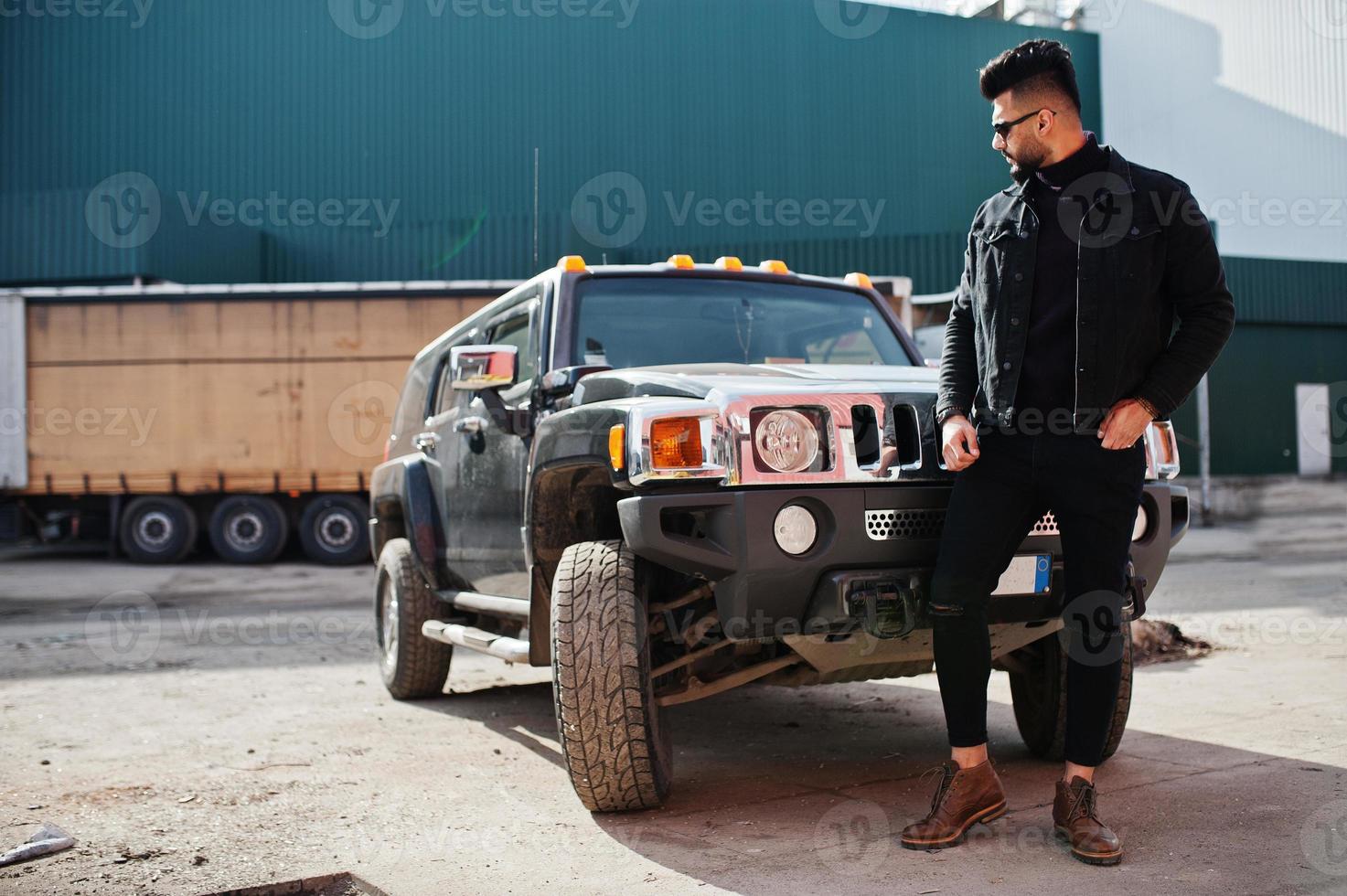 Fashion rich beard Arab man wear on black jeans jacket and sunglasses posed against big black suv car. Stylish, succesful and fashionable arabian model guy. photo