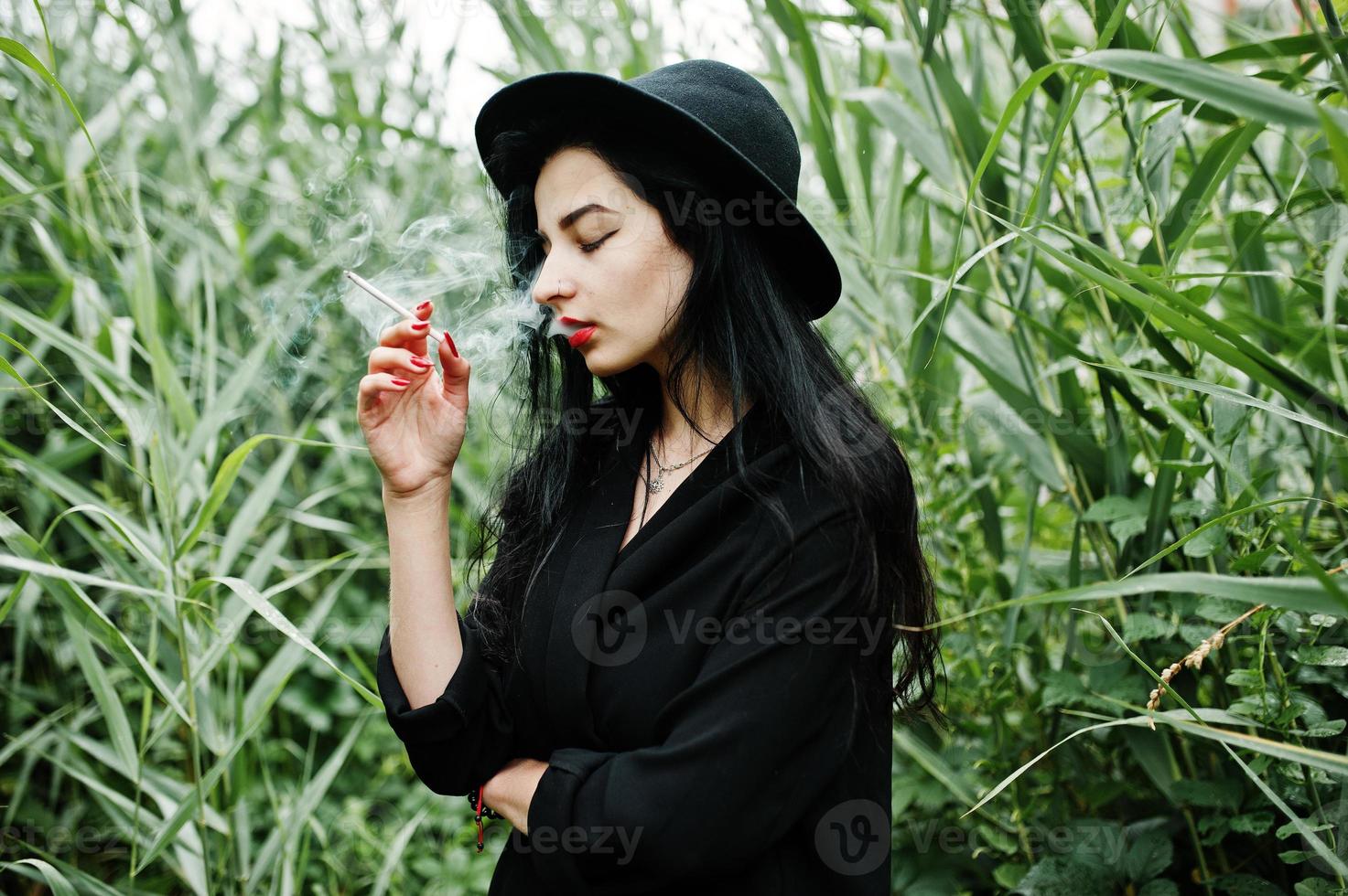 Sensual smoker girl all in black, red lips and hat. Goth dramatic woman smoking on common reed. photo