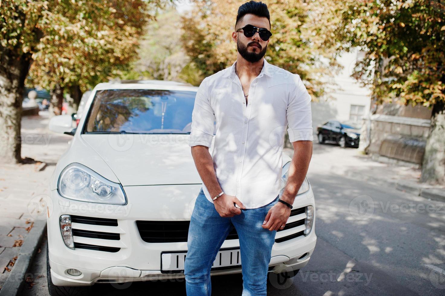 Stylish tall arabian man model in white shirt, jeans and sunglasses posed at street of city. Beard rich attractive arab guy against white suv car. photo