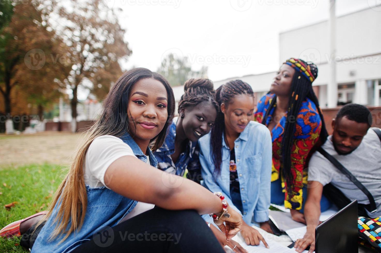 grupo de cinco estudiantes universitarios africanos que pasan tiempo juntos en el campus en el patio de la universidad. amigos afro negros sentados en el césped y estudiando con computadoras portátiles. foto