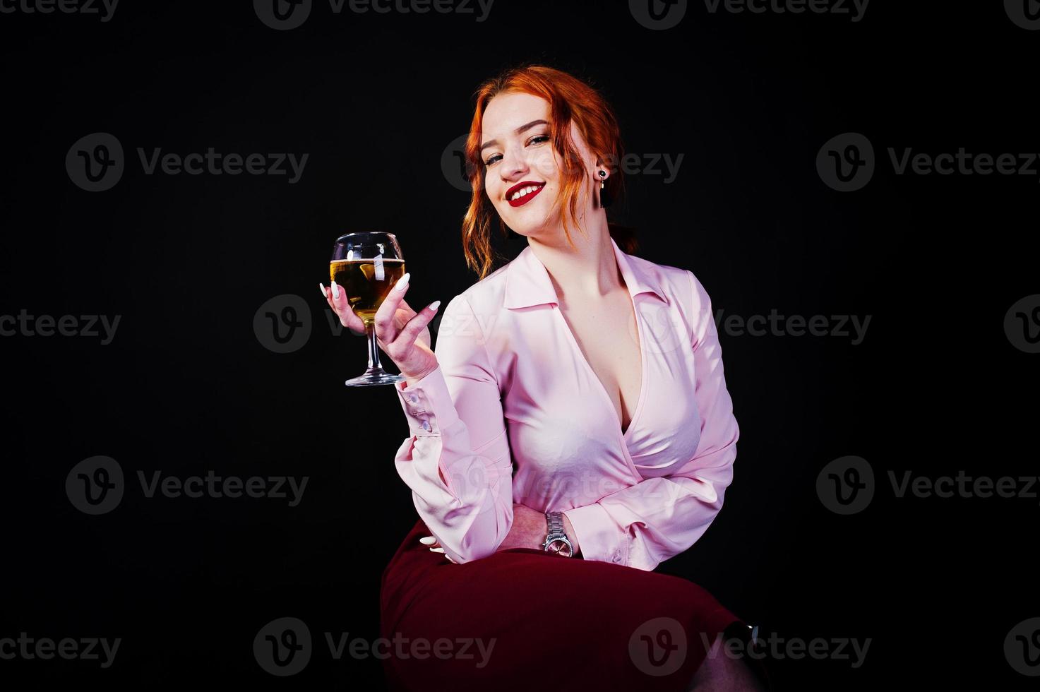 Gorgeous red haired girl in pink blouse and red skirt with glass of wine at hand isolated on black. photo
