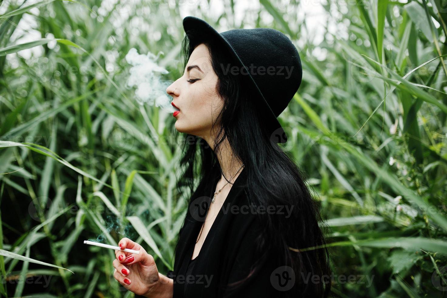 Sensual smoker girl all in black, red lips and hat. Goth dramatic woman smoking on common reed. photo