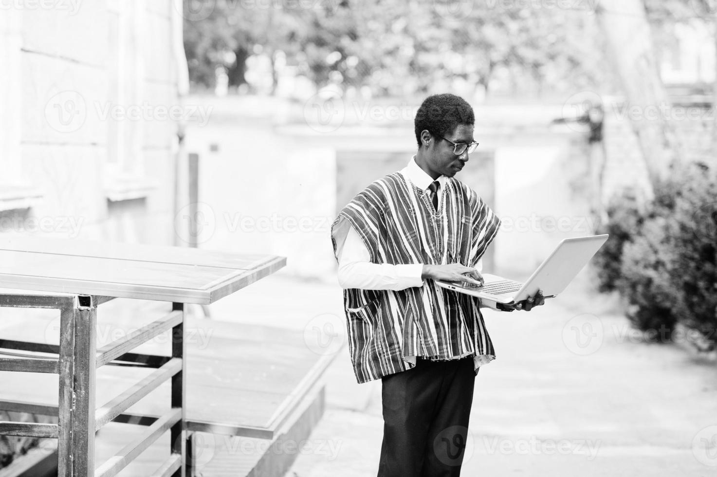 hombre africano con ropa tradicional y gafas con laptop trabajando al aire libre. foto