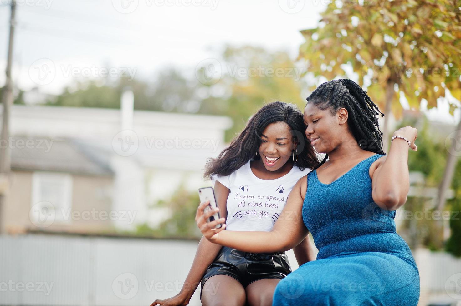 Two african american dark skinned friends female. One of them plus size model, second slim. Having fun and spending time together. Look at mobile phone. photo