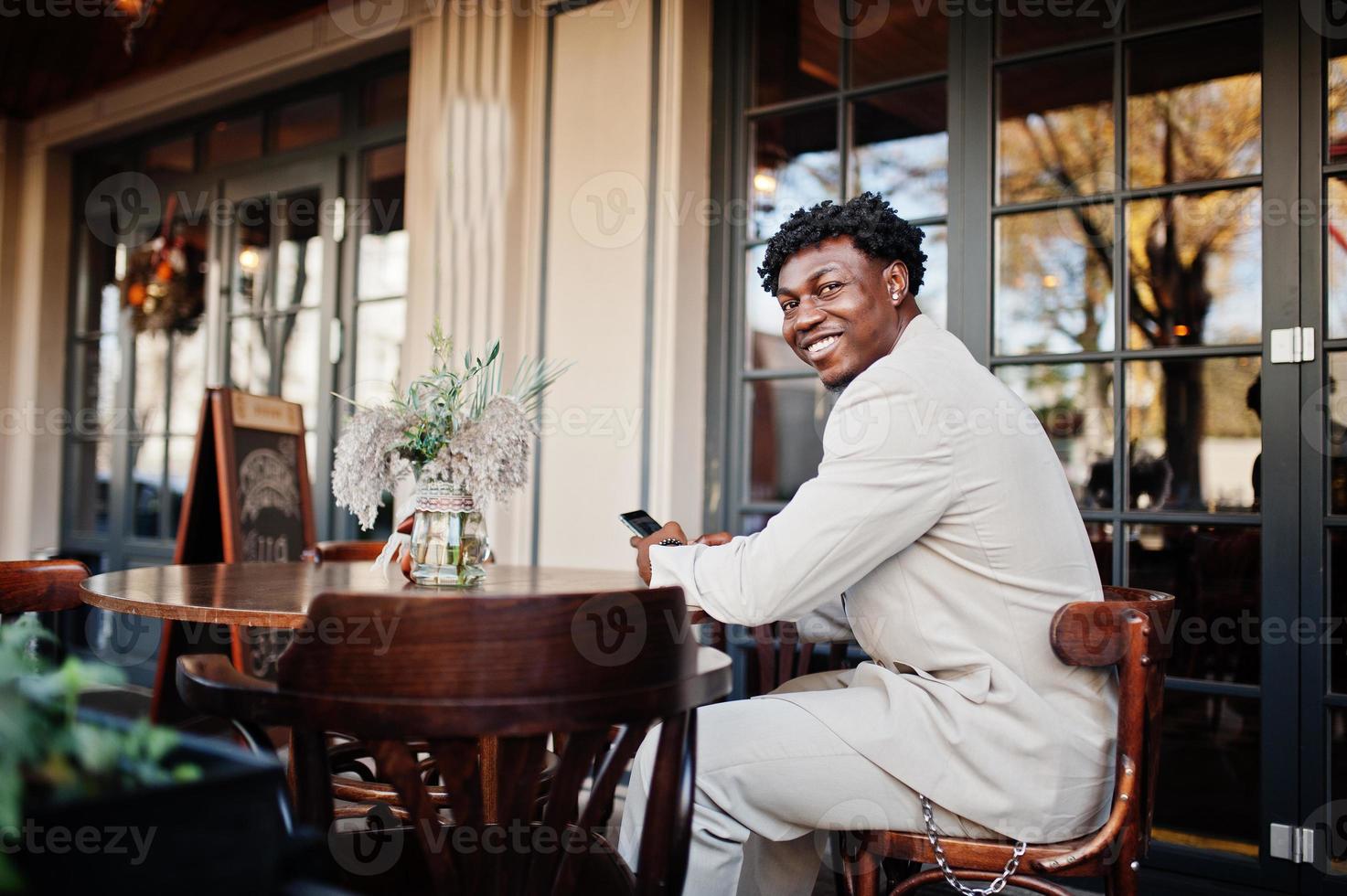 Stylish afro man in beige old school suit sitting on cafe with mobile phone. Fashionable young African male in casual jacket on bare torso. photo