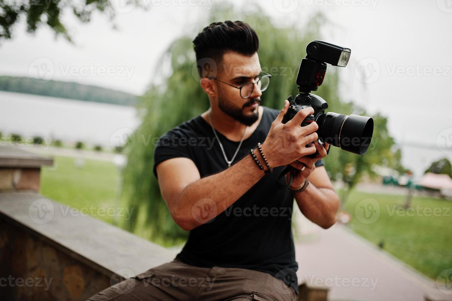 Impresionante y hermoso fotógrafo de hombre macho con barba árabe alta con gafas y camiseta negra con cámara profesional en las manos. foto