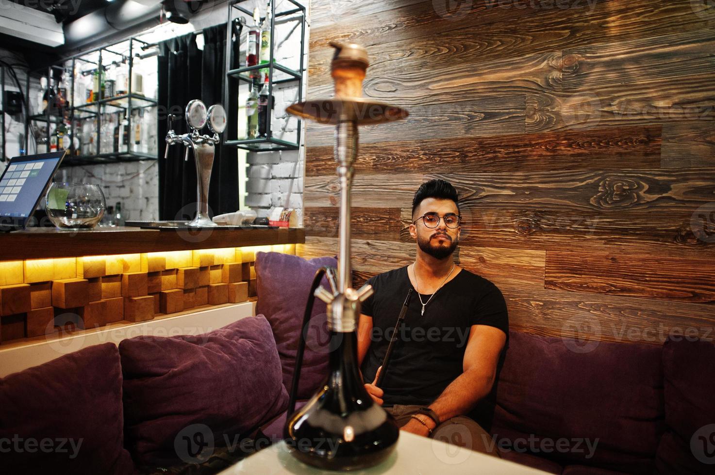 Stylish beard arabian man in glasses and black t-shirt smoking hookah indoor bar. Arab model having rest. photo