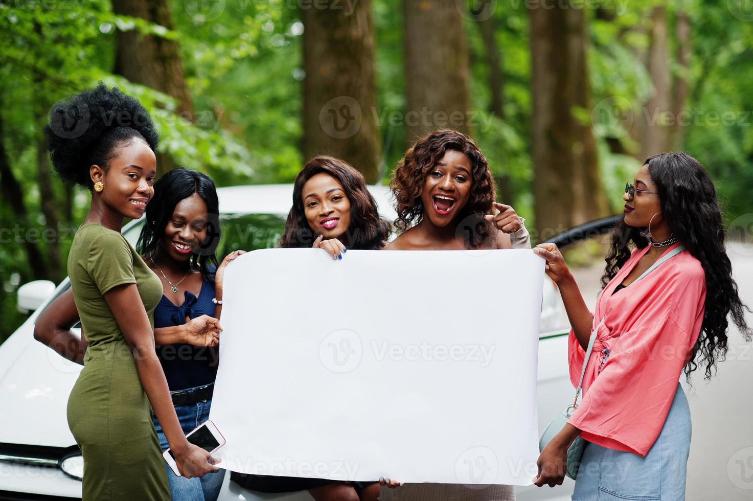 Group of five happy african american girls posed against car and hold empty white blank banner. Free space for your text. photo