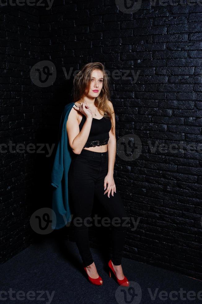 Handsome brunette girl wear on black and red high heels, posing at studio against dark brick wall. Studio model portrait. photo