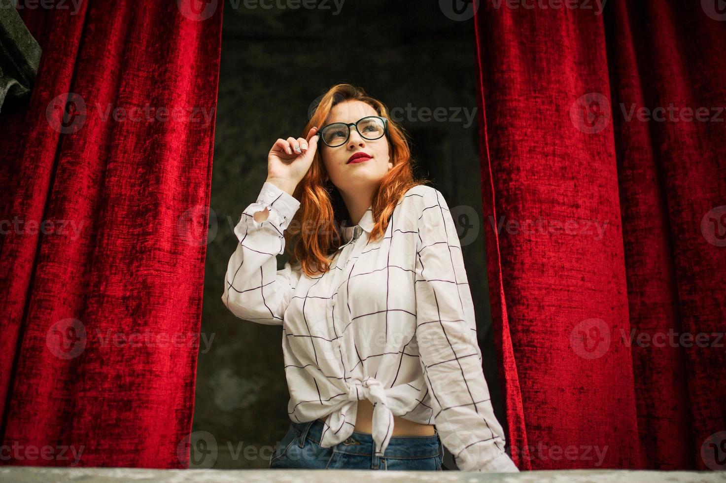 Attractive redhaired woman in eyeglasses, wear on white blouse posing at arch of open red curtains. photo