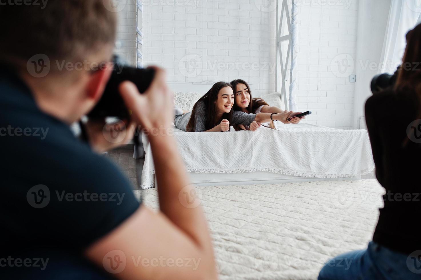 el equipo de dos fotógrafos disparando a gemelas modelos mientras hacen clic en el control remoto de la televisión en el estudio. fotógrafo profesional en el trabajo. foto