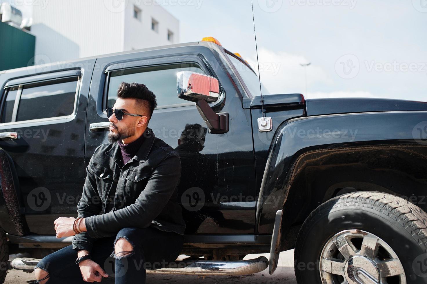 Fashion rich beard Arab man wear on black jeans jacket and sunglasses posed against big black suv car. Stylish, succesful and fashionable arabian model guy. photo