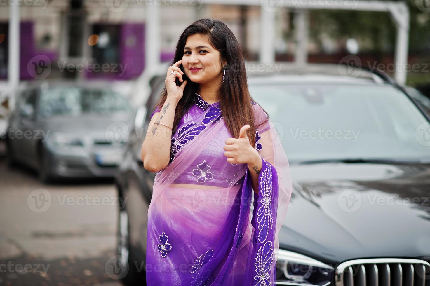 niña hindú india en el tradicional sari violeta posó en la calle contra un camión suv de negocios negro y hablando por teléfono móvil, muestra el pulgar hacia arriba. foto