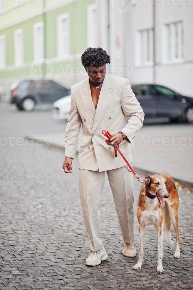 Stylish afro man in beige old school suit with Russian Borzoi dog. Fashionable young African male in casual jacket on bare torso. photo