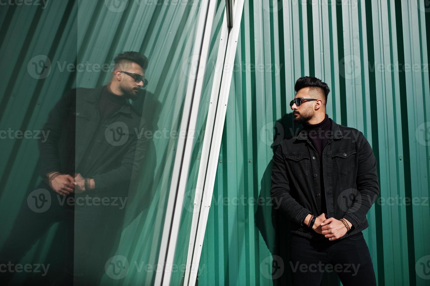 Fashion rich beard Arab man wear on black jeans jacket and sunglasses posed against green wall with windows. Stylish, succesful and fashionable arabian model guy. photo