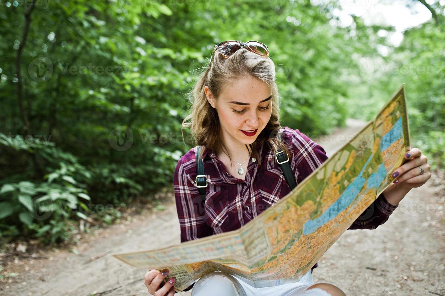 Portrait of a positive young gorgeous blonde sitting on the ground with a map in her hands in the forest. photo