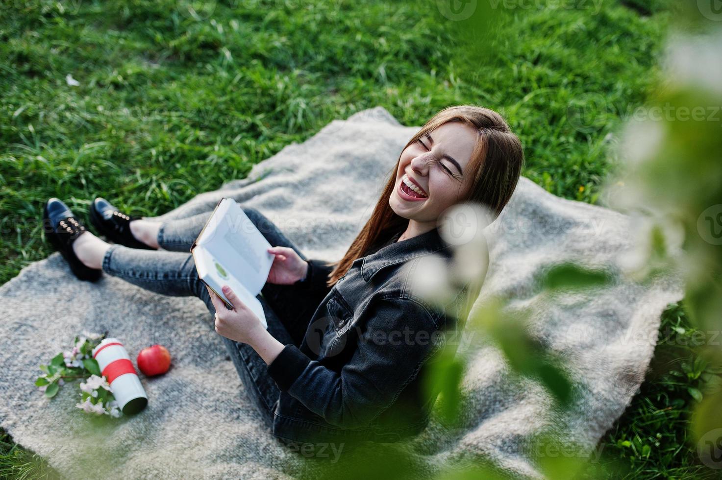 Young brunette girl at jeans sitting on plaid against spring blossom tree and read the book. photo