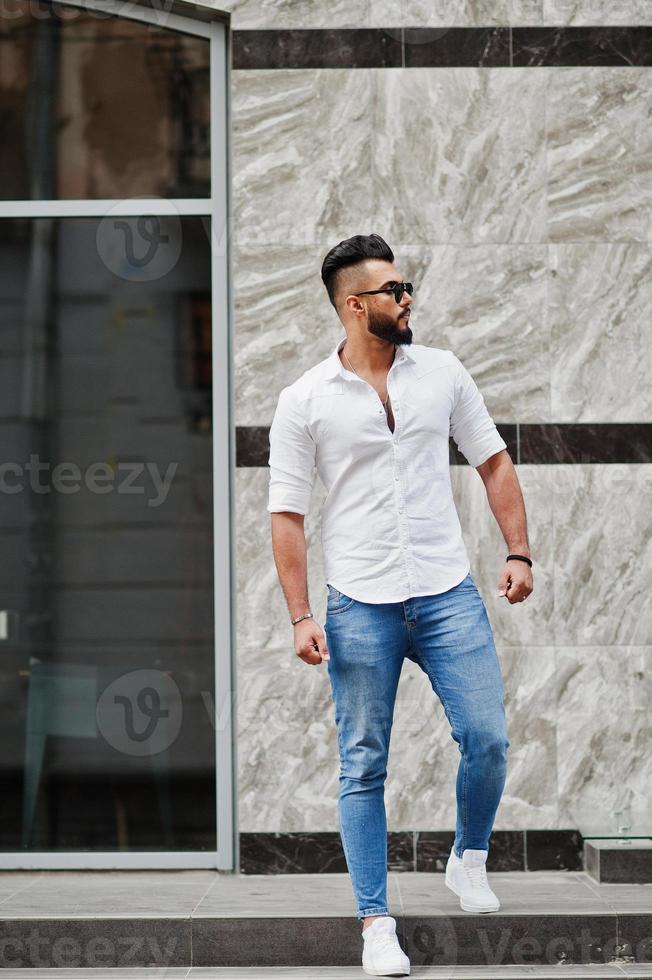 Elegante modelo de hombre alto con camisa blanca, jeans y gafas de sol en la calle de la ciudad. barba chico árabe atractivo. 10442407 Foto de stock en