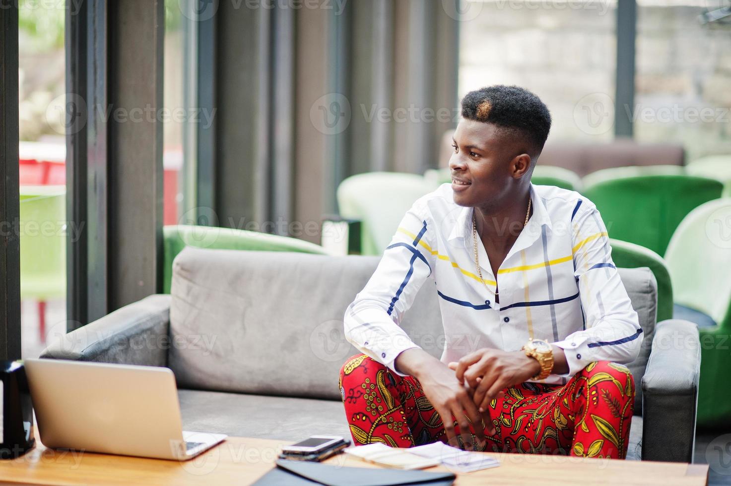 Portrait of handsome stylish african american business man in red throusers and white shirt sitting in office with laptop and cash on table. photo