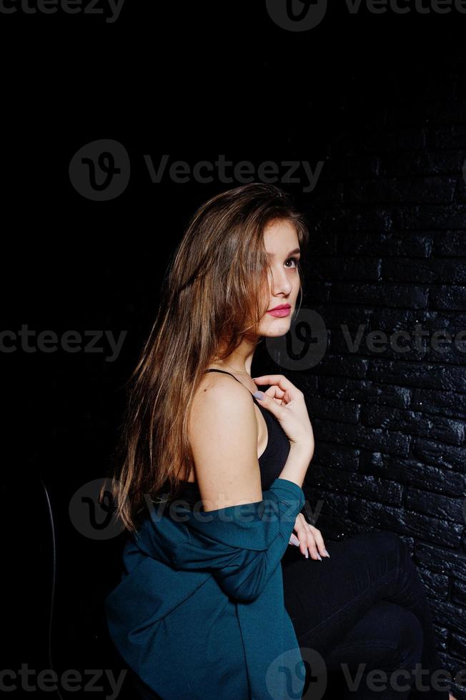 hermosa chica morena vestida con una chaqueta negra y verde, sentada y posando en una silla en el estudio contra una pared de ladrillo oscuro. retrato de modelo de estudio. foto