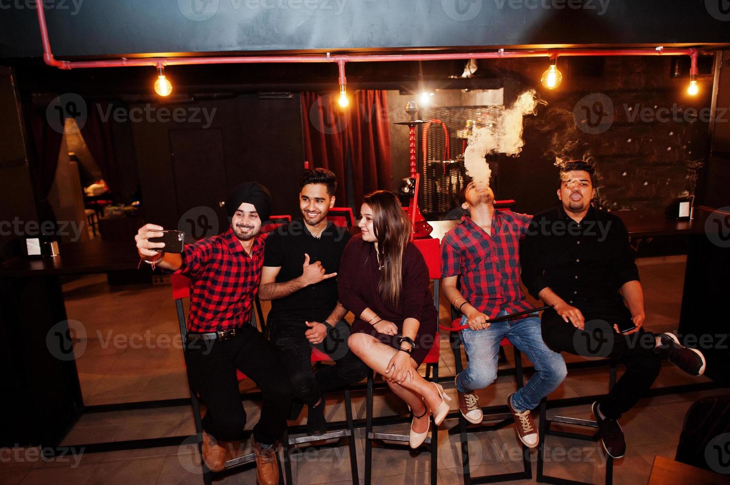 Group of indian friends sitting at lounge bar, having fun and rest, smoking hookah and looking on mobile phones to make selfie. photo