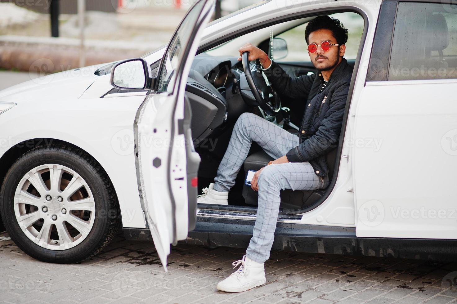 South asian man or indian male wear red eyeglasses sitting inside his white transportation car. photo