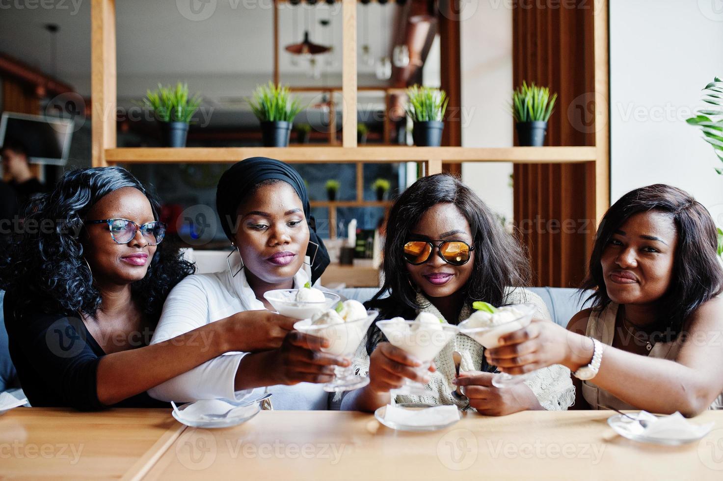 Four african american girls sitting on table at cafe and cheers with ice cream dessert. photo