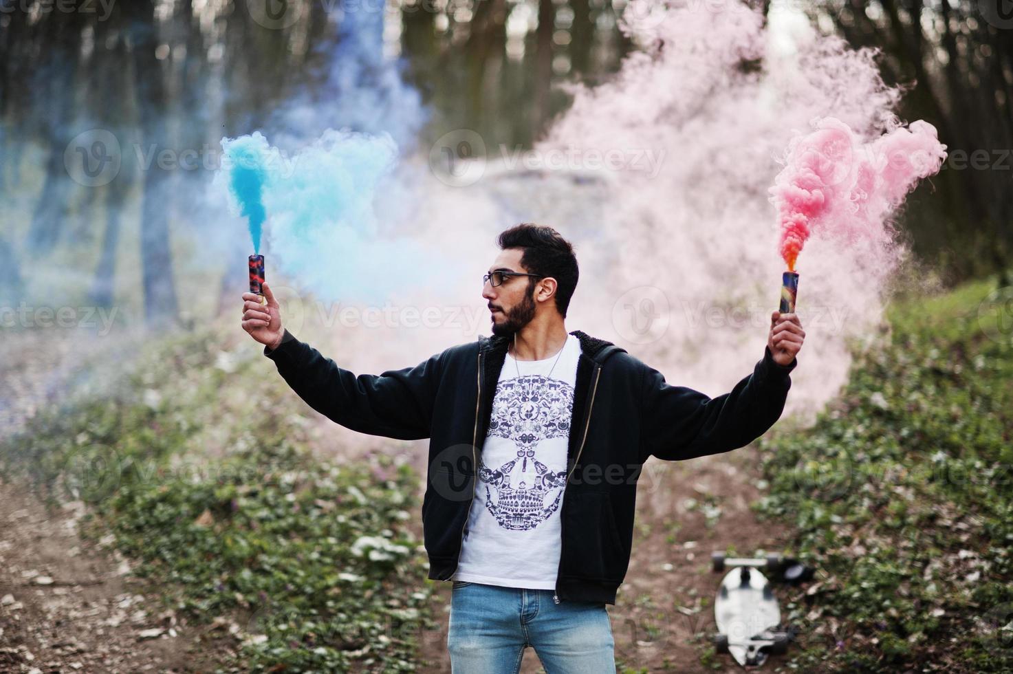 hombre árabe de estilo callejero con anteojos sostiene bengalas de mano con bomba de granada de humo rojo y azul. foto