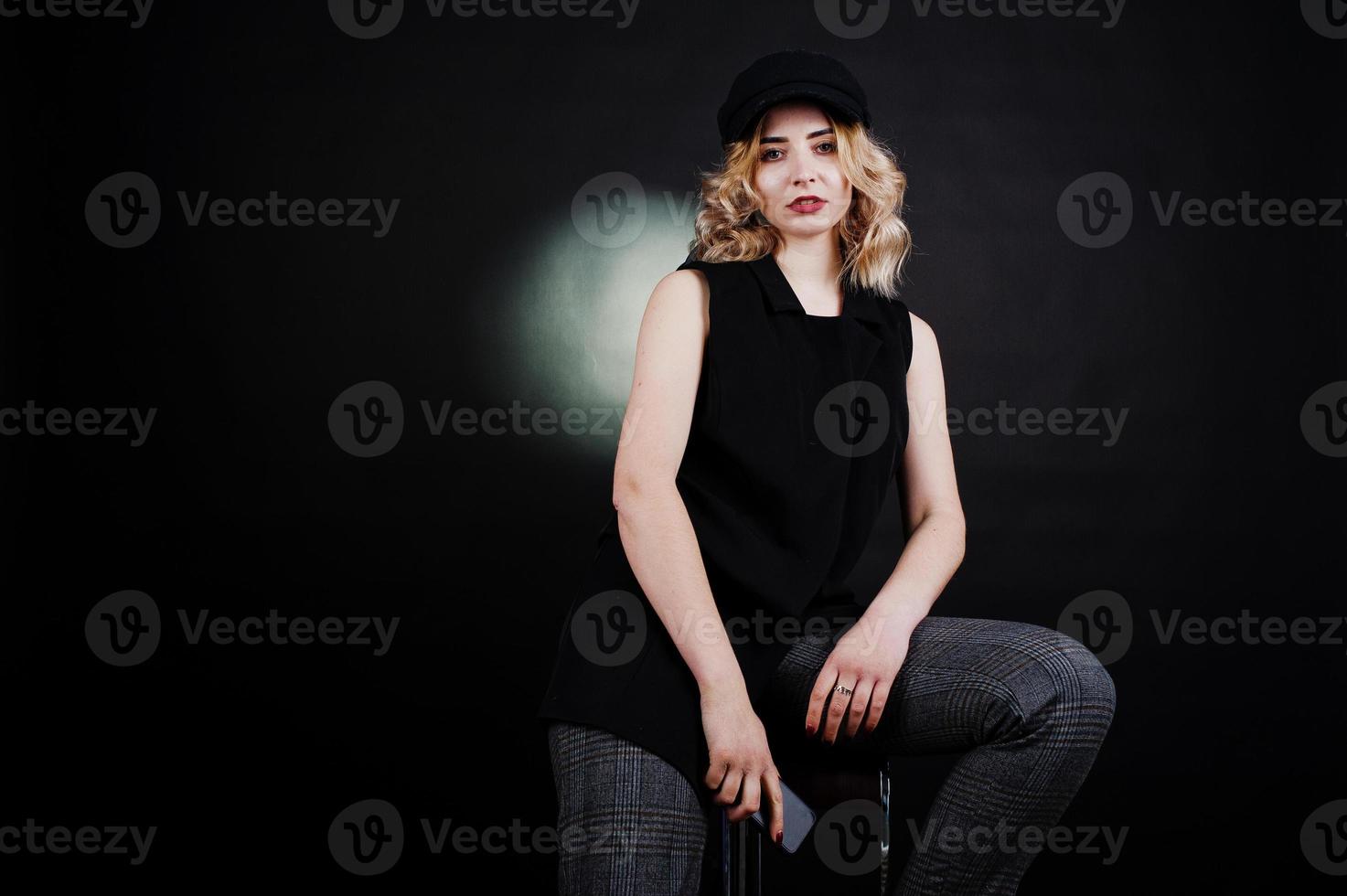 Studio portrait of blonde girl in black wear and cap with mobile phone at hand against dark background. photo