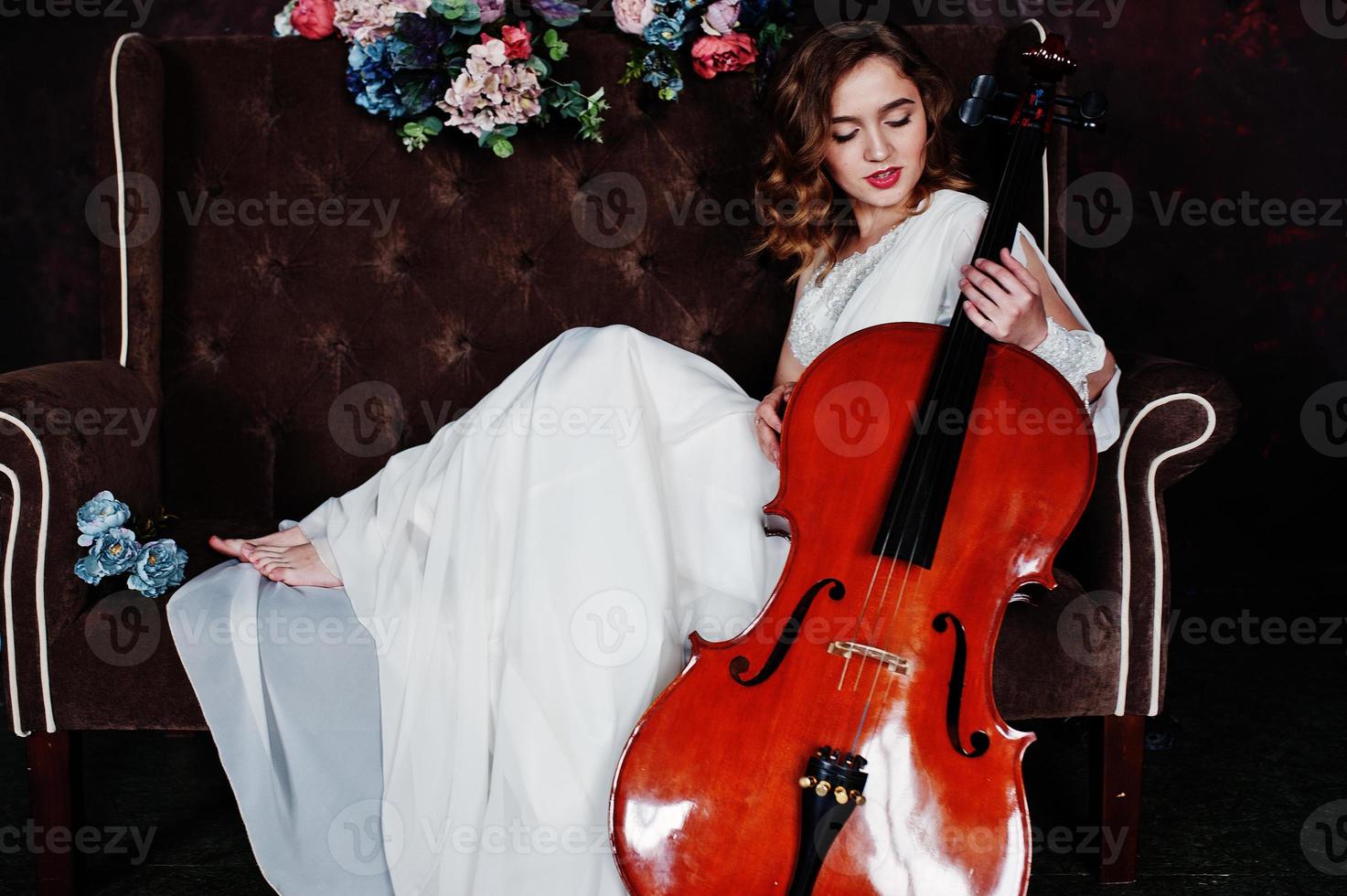 Pretty young gilrl musician in white dress with double bass sitting on brown vintage sofa. photo