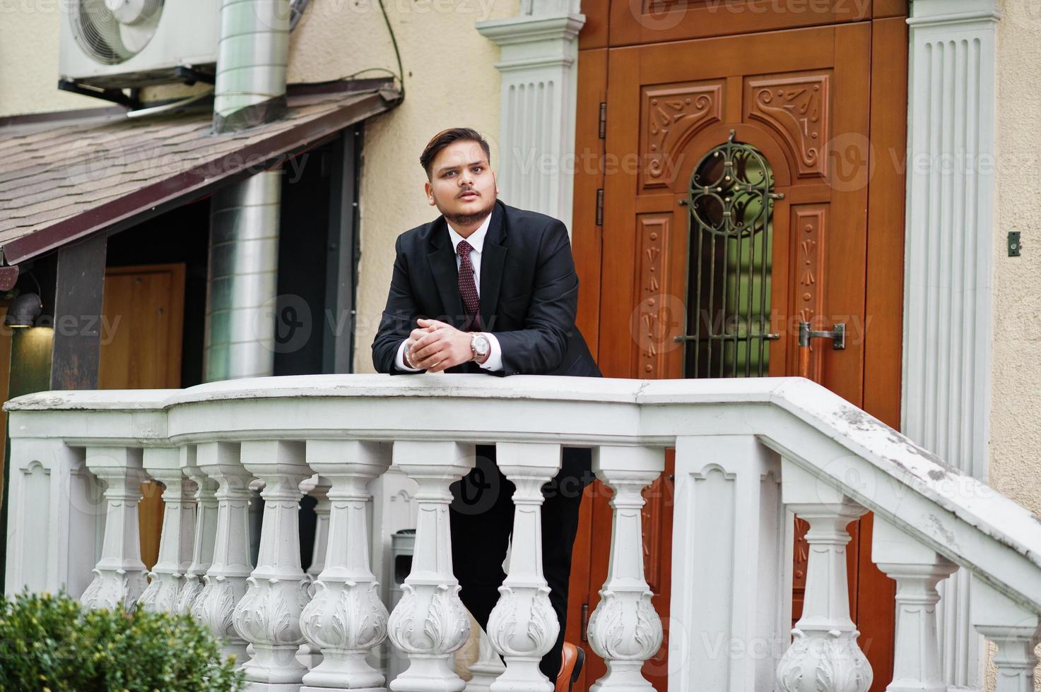 Stylish indian businessman in formal wear leaning on a railing against door in business center. photo