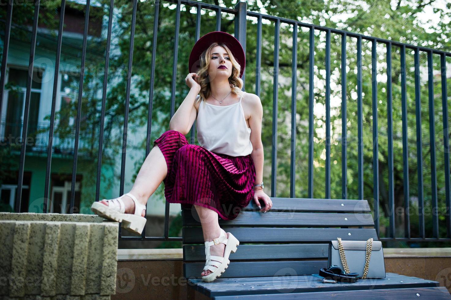 Fashionable and beautiful blonde model girl in stylish red velvet velour skirt, white blouse,hat and handbag, posed on bench. photo