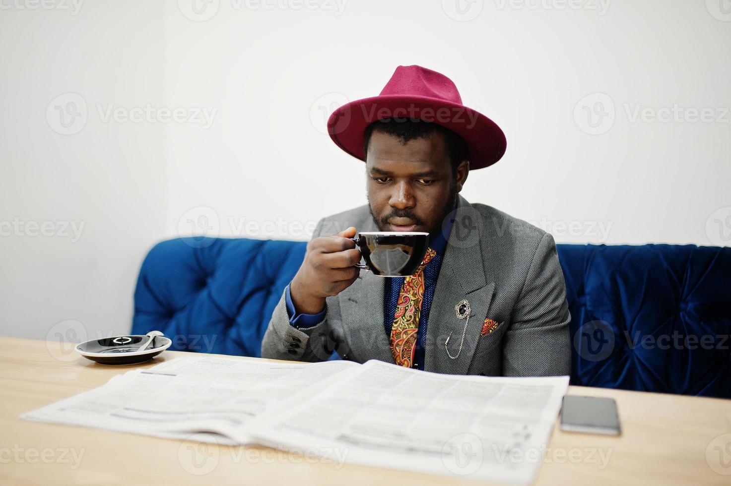 Stylish African American man model in gray jacket tie and red hat drink coffee at cafe and read newspapers. photo