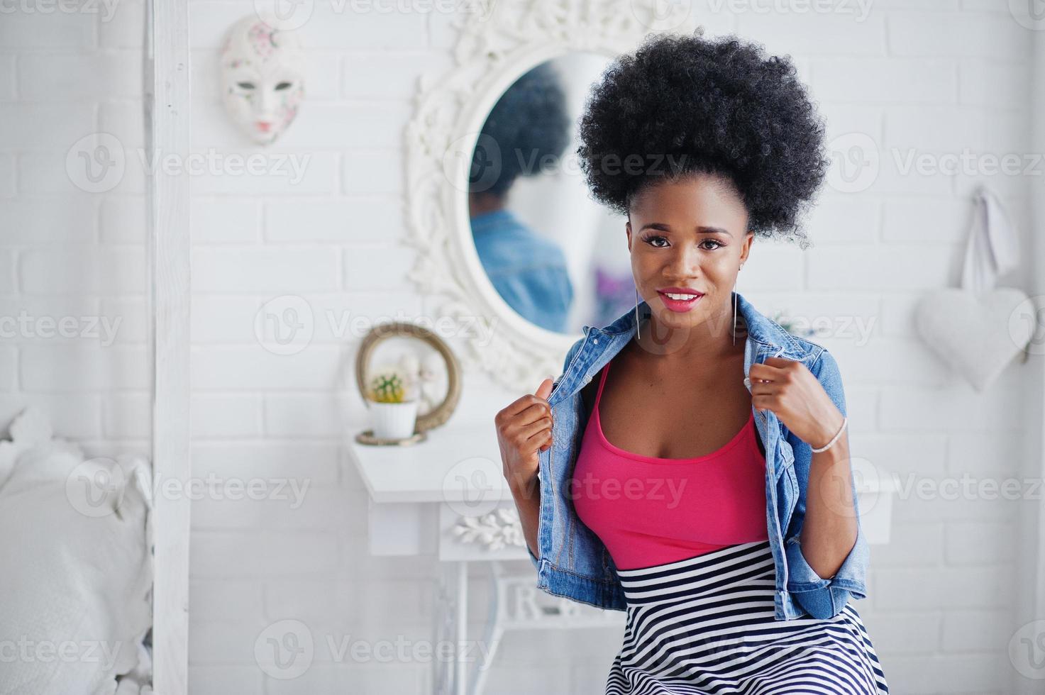 Attractive african american woman with afro hair wear on skirt and jeans jacket, posed at white room. Fashionable black model. photo