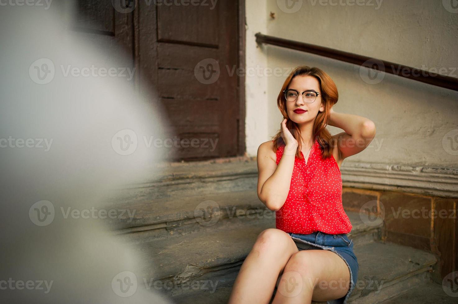 Attractive redhaired woman in eyeglasses, wear on red blouse and jeans skirt posing. photo