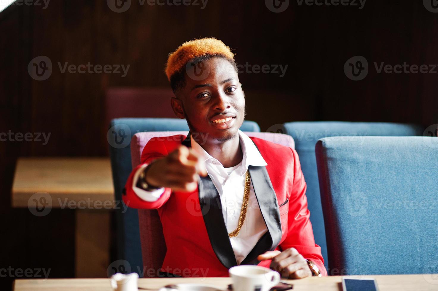 Fashion african american man model at red suit, with highlights hair sitting at cafe with coffee and show finger to you. photo
