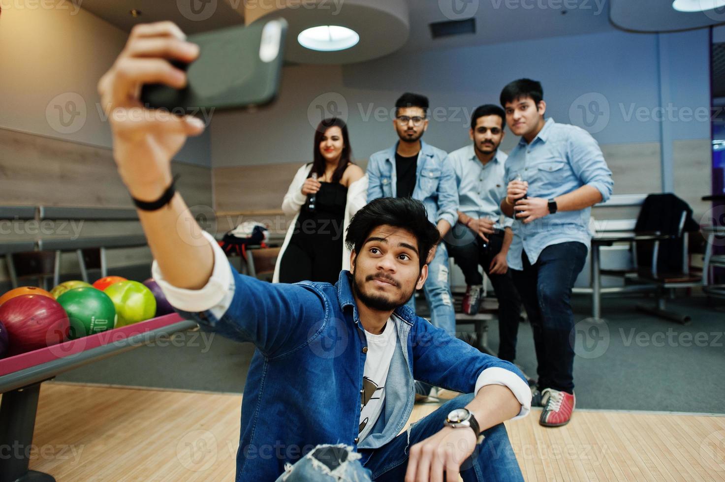 Group of five south asian peoples having rest and fun at bowling club. Making selfie by phone and holding cold soda drinks from glass bottles. photo