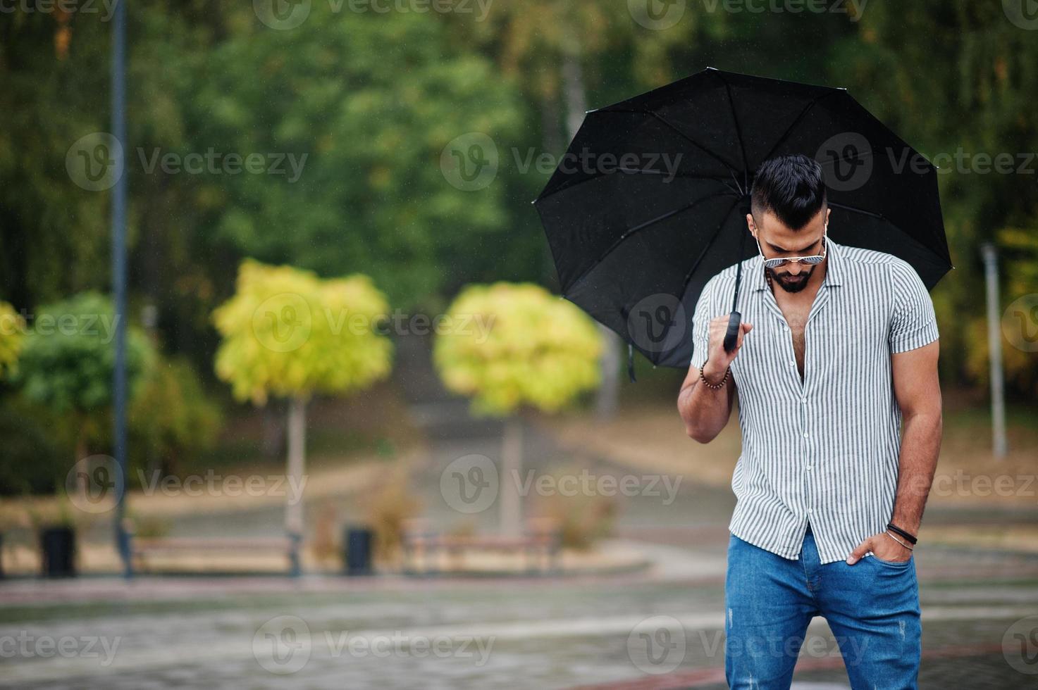 Fashionable tall arab beard man wear on shirt, jeans and sunglasses with umbrella posed at rain on park square. photo