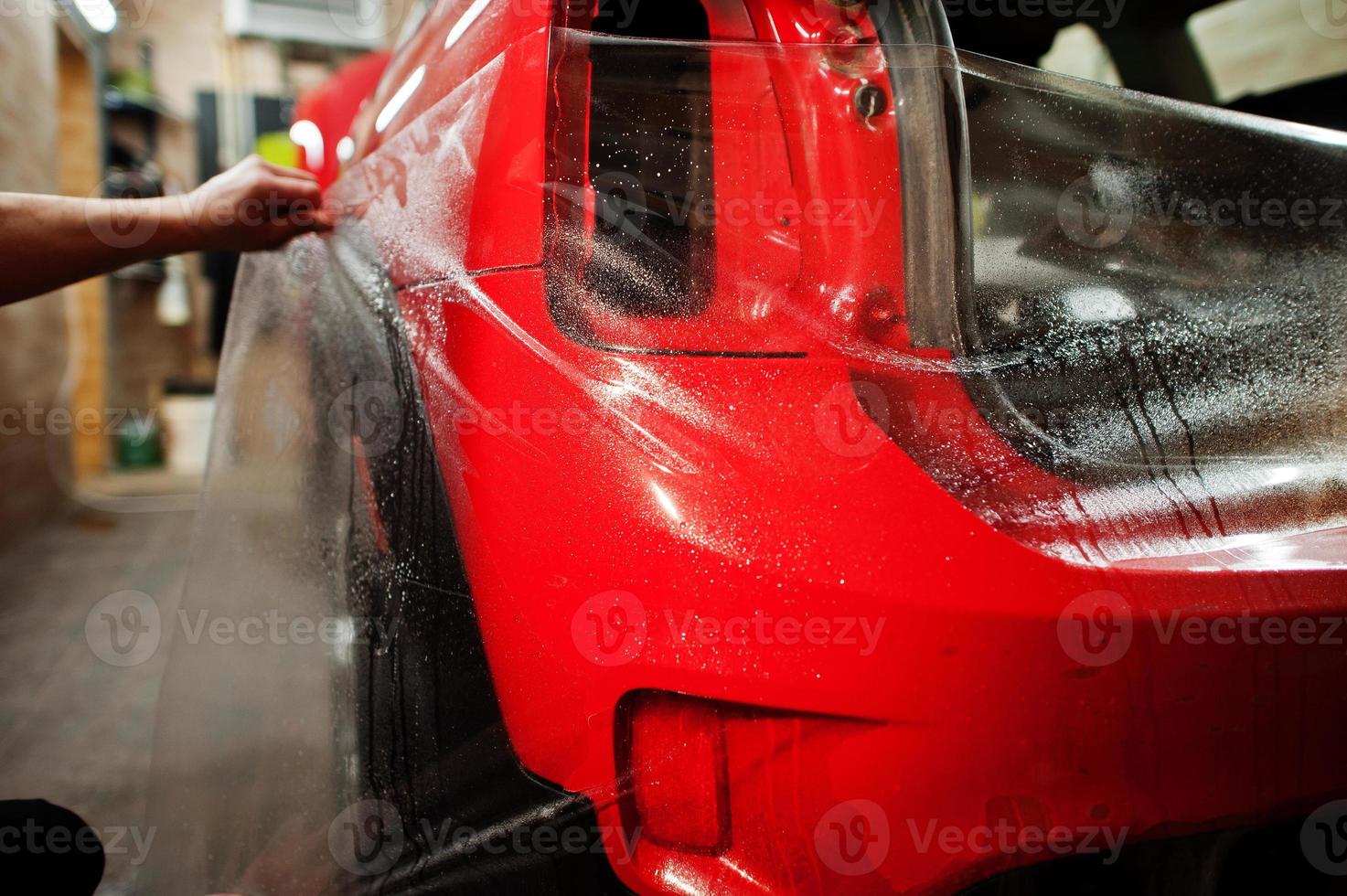 Car service worker put anti gravel film on a red car body at the detailing vehicle workshop. Car protection with special films. photo