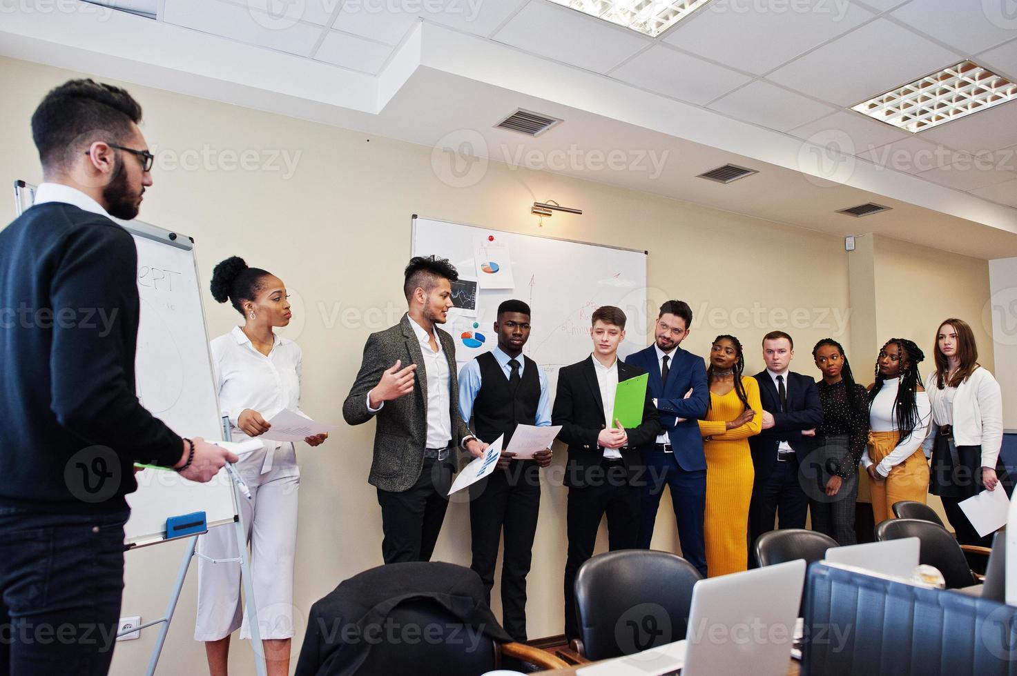 Mixed race business coach presenting report standing near whiteboard pointing on sales statistic shown on diagram and chart teach diverse company members gathered together in conference room. photo