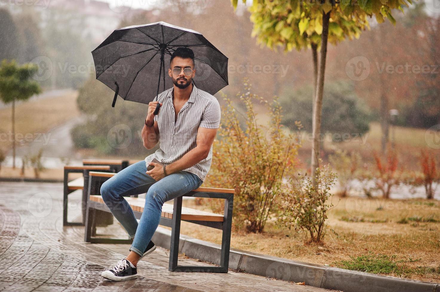 Fashionable tall arab beard man wear on shirt, jeans and sunglasses with umbrella posed at rain on park square. photo
