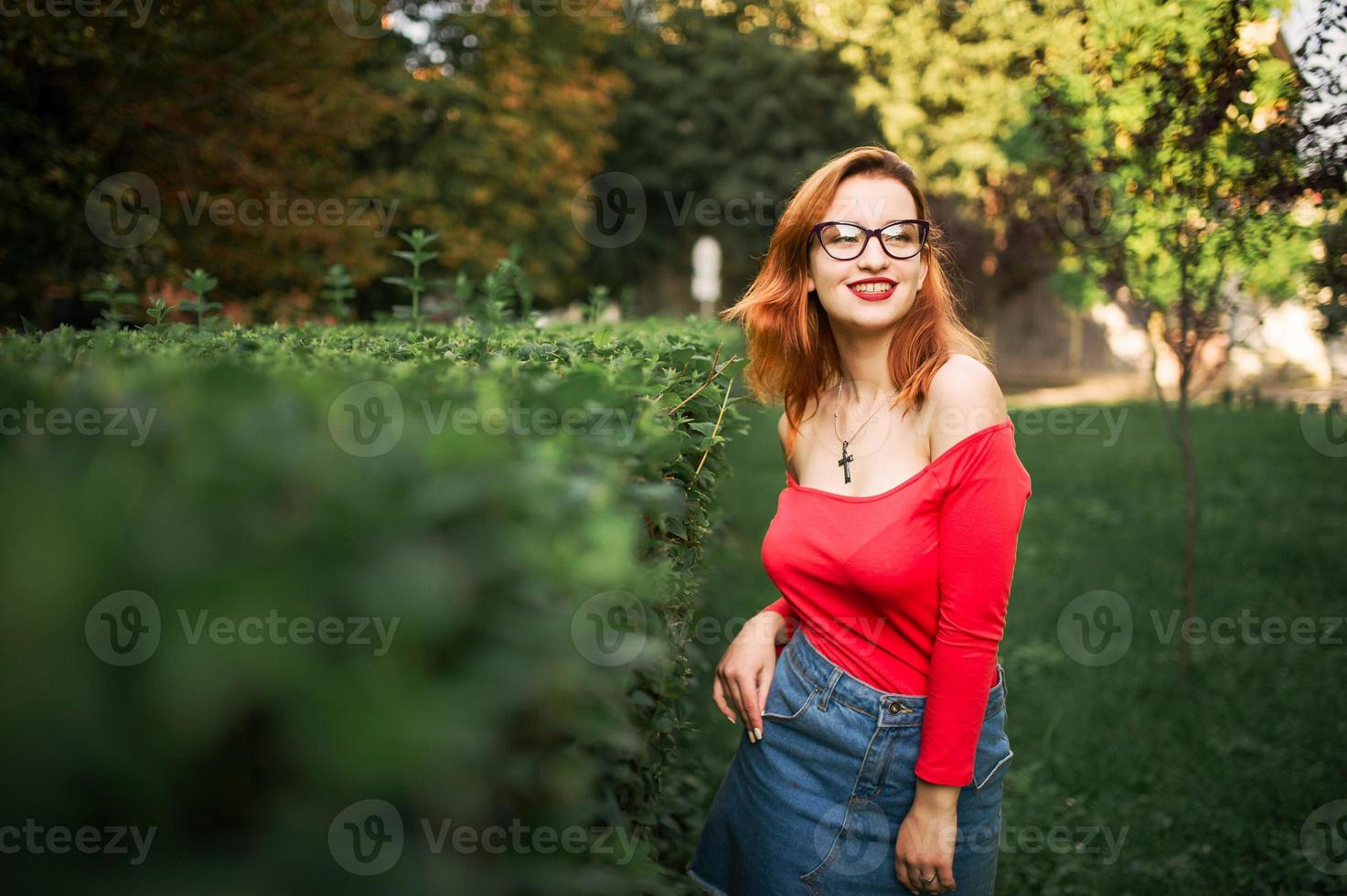 Attractive redhaired woman in eyeglasses, wear on red blouse and jeans skirt posing at green park. photo