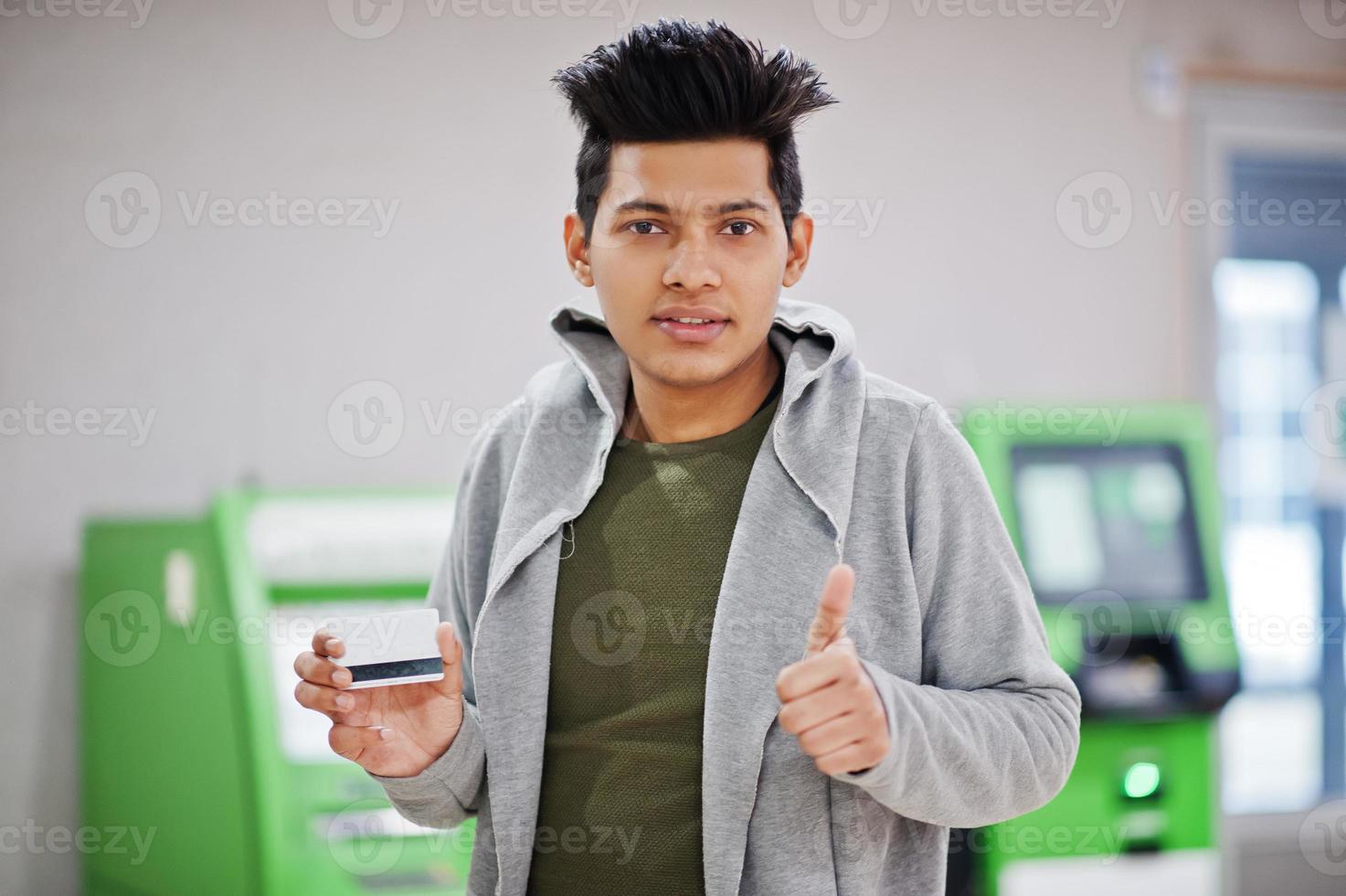 Young stylish asian man with credit card in hand show thumb up against row of green ATM. photo