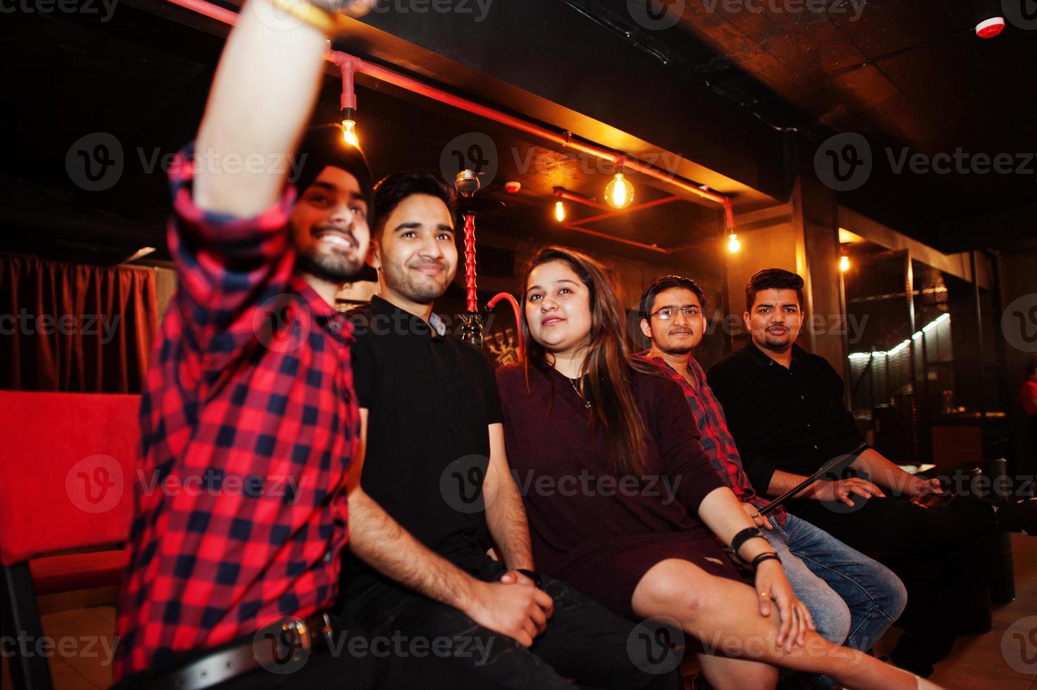 Group of indian friends sitting at lounge bar, having fun and rest, smoking hookah and looking on mobile phones to make selfie. photo