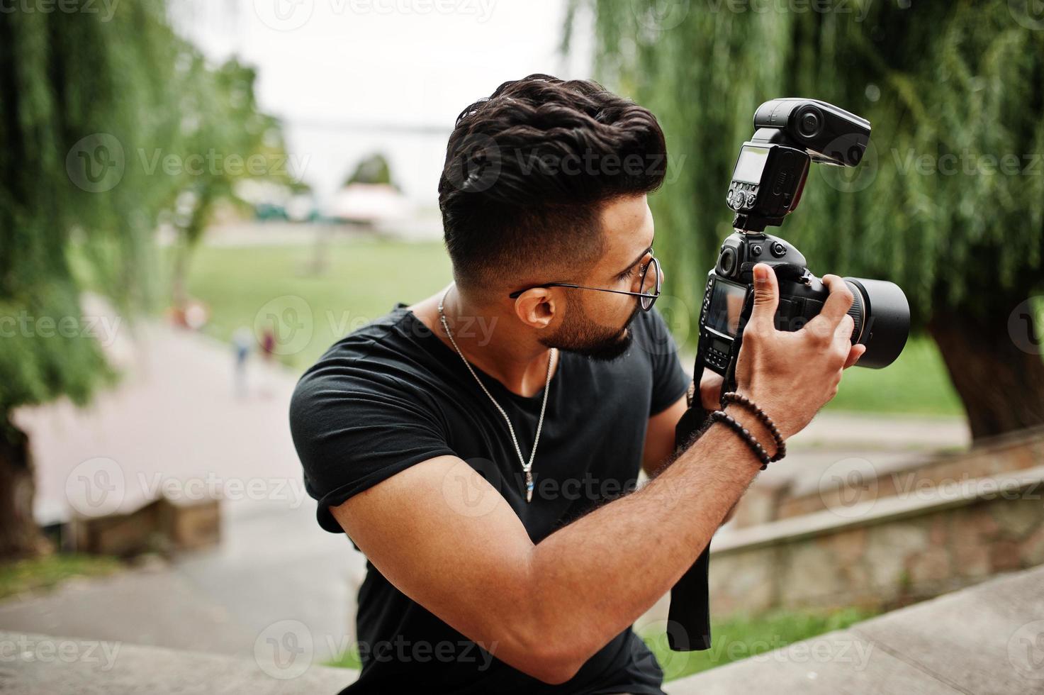 Impresionante y hermoso fotógrafo de hombre macho con barba árabe alta con gafas y camiseta negra con cámara profesional en las manos. foto